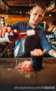 Male barman pours red beverage into the black glass full of ice. Barkeeper occupation, bartender job. Barman pours beverage into the glass full of ice