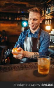 Male bartender in apron cleans orange at the bar counter. Alcohol beverage preparation. Barman occupation. Male bartender cleans orange at the bar counter