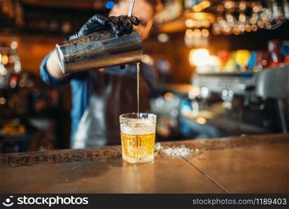 Male bartender in apron pours a drink through a sieve into a glass. Barman at the bar counter. Alcohol beverage preparation. Bartender pours drink through sieve into a glass