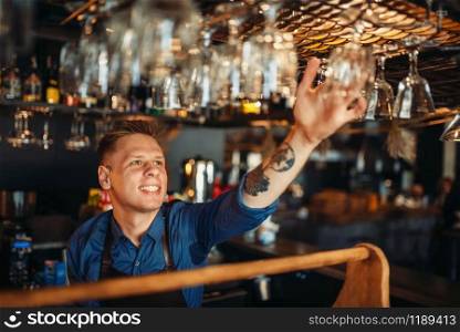 Male bartender in apron takes a clean glass from bar counter. Barman occupation, bartending. Male bartender takes clean glass from bar counter