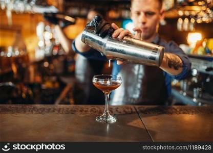 Male bartender pouring the drink from the shaker at the bar counter. Alcoholic coctail preparation. Barman occupation, bartending. Male bartender pouring drink from the shaker