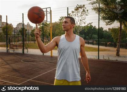 Male basketball player spinning the ball on his finger on outdoor court. Male athlete in sportswear on streetball training, summer stadium