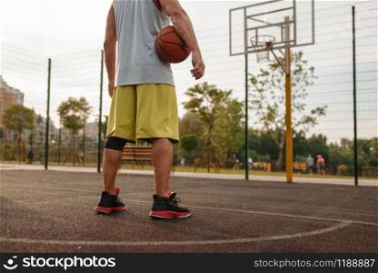 Male basketball player with ball standing at the basket on outdoor court, back view. Male athlete in sportswear on streetball training, summer stadium