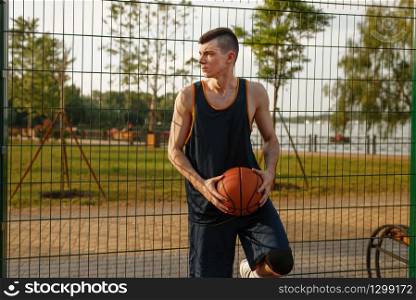 Male basketball player with ball standing at the mesh fence on outdoor court, side view. Male athlete in sportswear on streetball training, summer stadium. Basketball player standing at the mesh fence