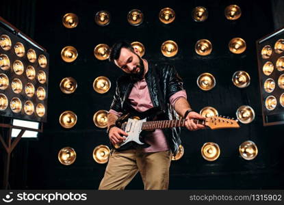 Male bearded guitarist in black leather jacket on the stage with the decorations of lights. Performer with electro guitar, solo concert