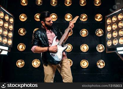 Male bearded guitarist in black leather jacket on the stage with the decorations of lights. Performer with electro guitar, solo concert. Male guitarist on stage with decorations of lights
