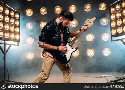 Male bearded guitarist in black leather jacket on the stage with the decorations of lights. Performer with electro guitar, solo concert. Male guitarist on stage with decorations of lights