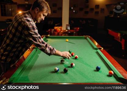 Male billiard player places balls on green table, poolroom interior on background. Man plays american pool game in sport bar