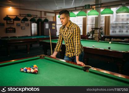 Male billiard player with cue at the green table with colorful balls, poolroom interior on background. Man plays american pool game in sport bar