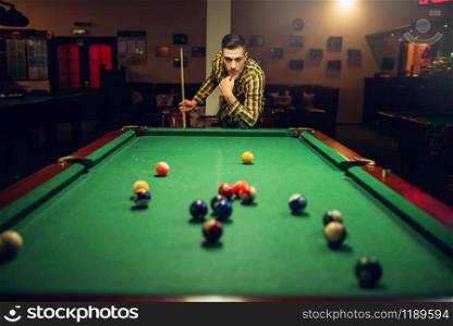 Male billiard player with cue thinking how to hit at the table with colorful balls. Man plays american pool game in sport bar