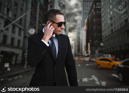 Male bodyguard in suit and sunglasses talking by security earpiece, chaos on city street on background, safe control. Professional guarding is a risky profession