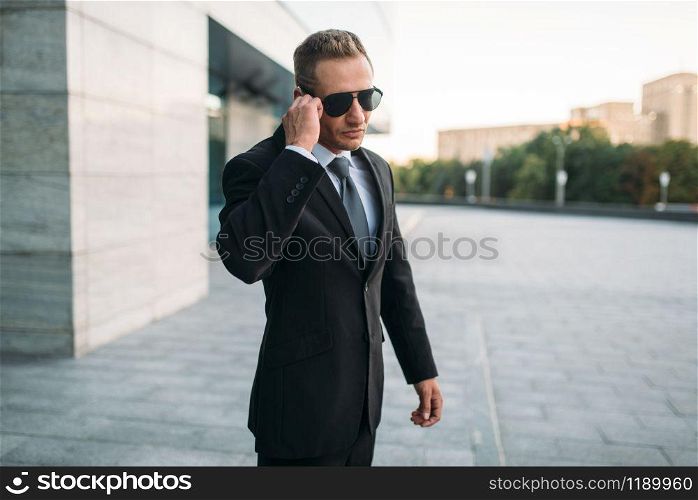 Male bodyguard in suit and sunglasses talking by security earpiece outdoors. Professional guarding is a risky profession