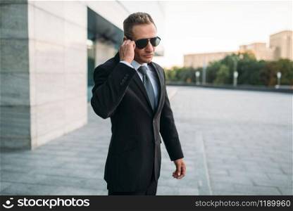Male bodyguard in suit and sunglasses talking by security earpiece outdoors. Professional guarding is a risky profession