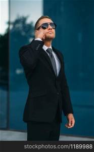 Male bodyguard talking by earpiece outdoors, security communication tools. Professional guarding is a risky profession. Guard in suit and sunglasses. Bodyguard talking by earpiece, communication tools