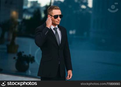 Male bodyguard uses security earpiece outdoors, professional communication tools. Guarding is a risky profession. Guard in suit and sunglasses. Male bodyguard uses security earpiece outdoors