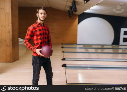 Male bowler standing on lane and holds ball in hands, back view. Bowling alley player poses in club, active leisure