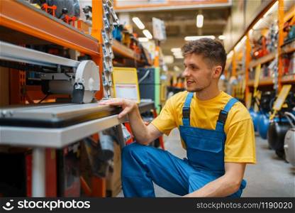 Male builder choosing circular saw at the shelf in hardware store. Constructor in uniform look at the goods in diy shop