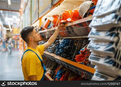 Male builder choosing helmet at the shelf in hardware store. Constructor in uniform look at the goods in diy shop