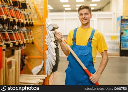 Male builder choosing sledgehammer at the shelf in hardware store. Constructor in uniform look at the goods in diy shop