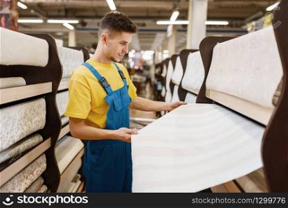 Male builder choosing wallpapers in hardware store. Constructor in uniform look at the goods in diy shop