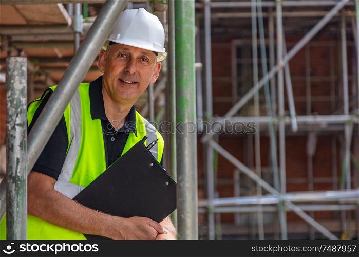 Male builder foreman, construction worker, engineer, surveyor or site manager holding a clipboard, wearing a white hard hat and hi vis vest standing on scaffolding on building site