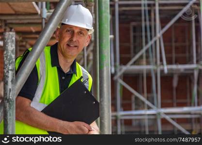 Male builder foreman, construction worker, engineer, surveyor or site manager holding a clipboard, wearing a white hard hat and hi vis vest standing on scaffolding on building site