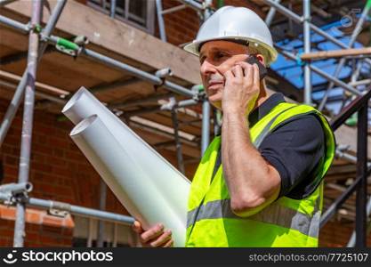 Male builder foreman, worker or architect on construction site holding building plans and talking on his cell phone