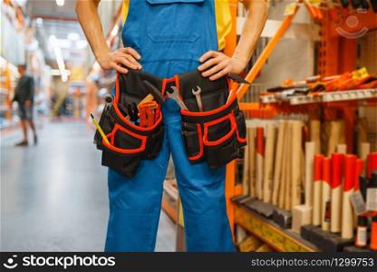 Male builder trying on tool belt at the shelf in hardware store. Constructor in uniform look at the goods in diy shop. Male builder trying on tool belt in hardware store
