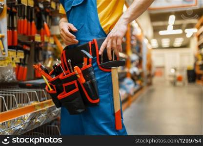 Male builder trying on tool belt at the shelf in hardware store. Constructor in uniform look at the goods in diy shop