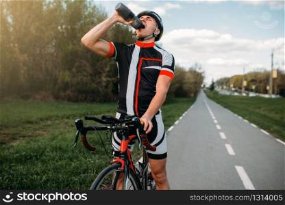 Male bycyclist in helmet and sportswear drinks water while training. Workout on bike path, cycling on asphalt road