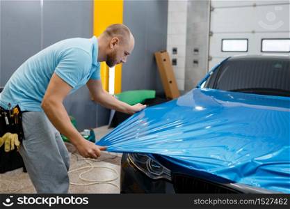 Male car wrapper puts protective vinyl foil or film on hood. Worker makes auto detailing. Automobile paint protection coating, professional tuning. Car wrapper puts protective foil or film on hood