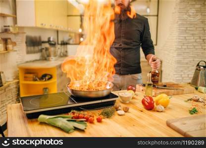 Male chef cooking meat with vetables on fire, flame into the frying pan, kitchen on background. Man preparing beef on table electric stove