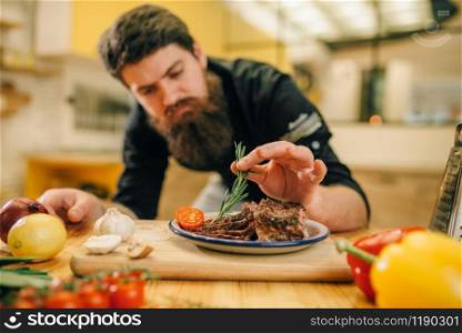 Male chef decorates with herbs roasted meat slices in a plate, kitchen on background. Man preparing beef with vegetables on countertop