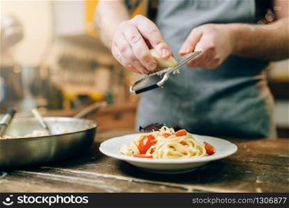 Male chef grates cheese in to the plate with fresh cooked pasta, pan on wooden kitchen table. Homemade fettuccine preparation process. Traditional italian cuisine