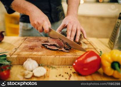Male chef hands with knife cuts roasted meat on slices, top view. Man preparing beef with vegetables. Chef at the pan with roasted meat and vetables
