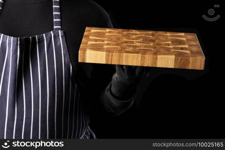 male chef in a striped blue apron and black clothes stands against a black background and holds in his hand a rectangular wooden kitchen chopping board, close up