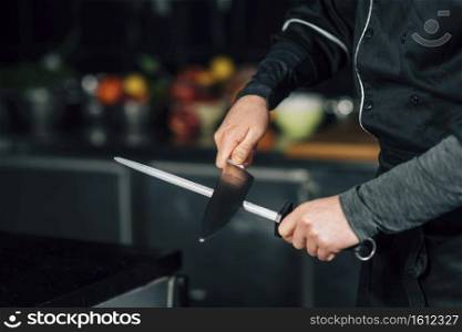 Male chef in black uniform sharpening the knives in the kitchen.