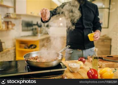 Male chef salted meat with vetables into the frying pan on the kitchen. Man preparing beef on table electric stove