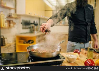 Male chef salted meat with vetables into the frying pan on the kitchen. Man preparing beef on table electric stove. Chef salted meat with vetables into the frying pan