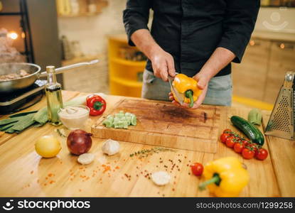 Male chef with knife cuts yellow pepper on wooden board, top view. Man cutting vegetables, fresh salad cooking, kitchen interior on background. Chef with knife cuts yellow pepper on wooden board