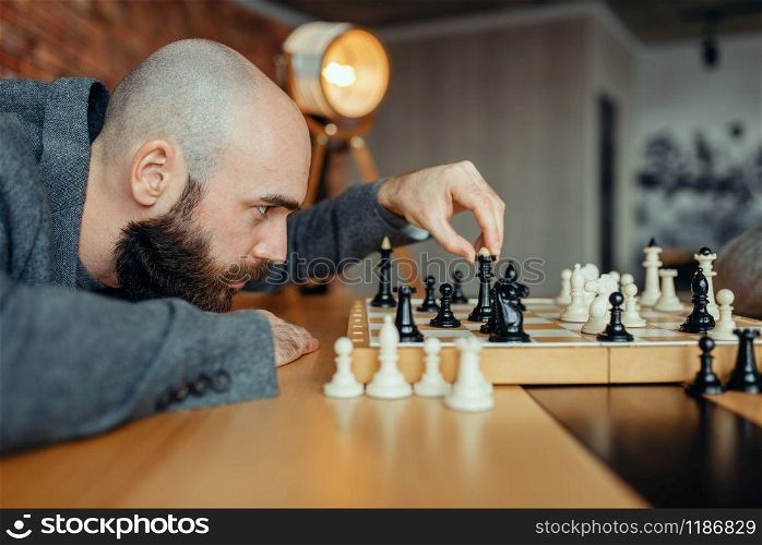 Male chess player playing black figures, queen move. Chessplayer at board, intellectual tournament indoors. Chessboard on wooden table, strategy game