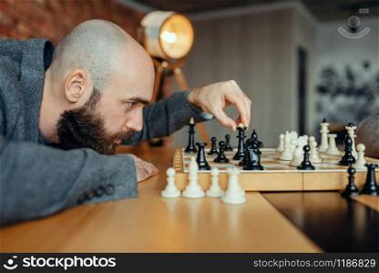 Male chess player playing black figures, queen move. Chessplayer at board, intellectual tournament indoors. Chessboard on wooden table, strategy game