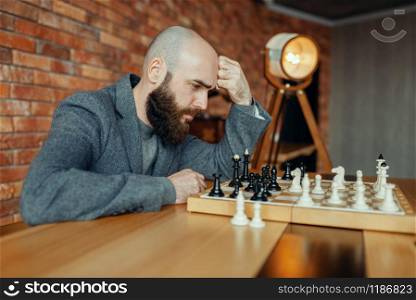 Male chess player playing black figures, thinking process. Chessplayer at board, intellectual tournament indoors. Chessboard on wooden table, strategy game. Male chess player playing, thinking process