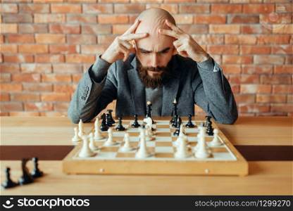 Male chess player playing black figures, thinking process. Chessplayer at board, intellectual tournament indoors. Chessboard on wooden table, strategy game