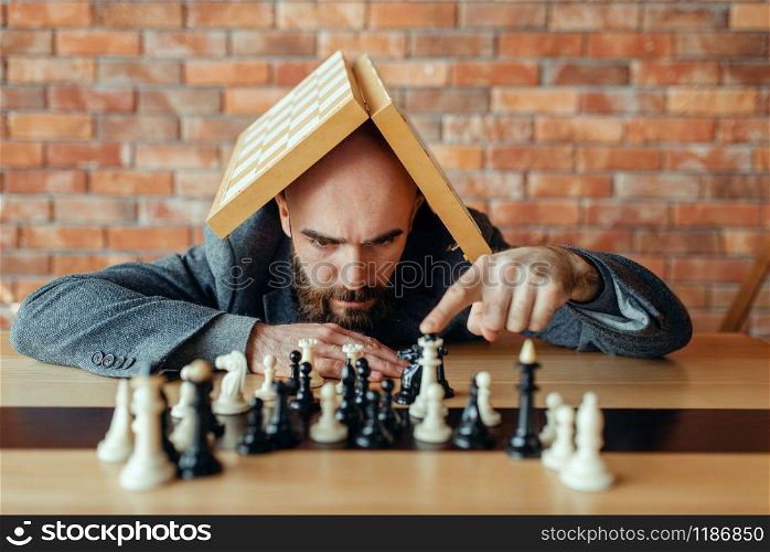 Male chess player with board on his head looking on figures, thinking process. Chessplayer playing on intellectual tournament indoors. Chessboard on wooden table, strategy game