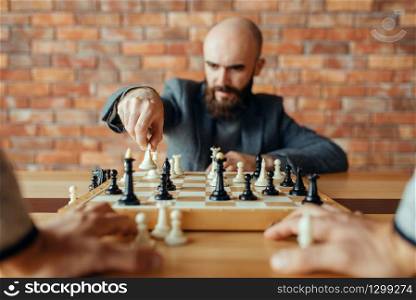 Male chess player with white figure in hand, queen move. Chessplayer at board, front view, intellectual tournament indoors. Chessboard on wooden table, strategy game