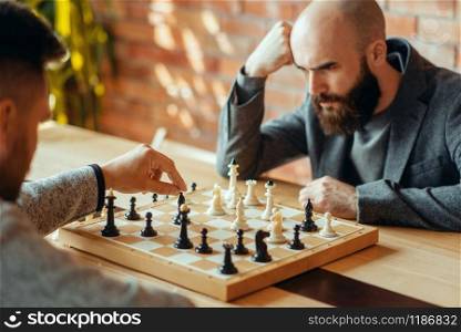 Male chess players playing at board, move the black elephant. Two chessplayers begin the intellectual tournament indoors. Chessboard on wooden table. Male chess players, move the black elephant