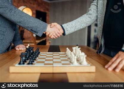 Male chess players shake hands before the game. Two chessplayers begin the intellectual tournament indoors. Chessboard on the table. Male chess players shake hands before the game