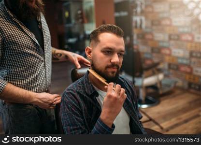 Male client combing his beard at the barbershop, professional barber on background