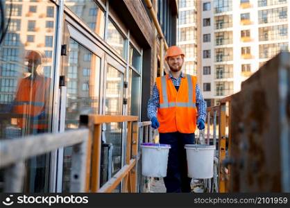 Male construction worker carrying buckets of cement, plaster or paint walking on scaffold. Male construction worker carrying buckets of cement